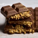 A close-up image of a rich, dark chocolate bar with a delectable filling of caramel and shredded coconut. The chocolate is smooth and glossy, with a hint of gold dust adding a touch of luxury. This is a Dubai chocolate bar, known for its exquisite combination of flavors and textures.