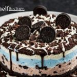 A layered Oreo ice cream cake with blue and pink ice cream layers, topped with chocolate drizzle, chocolate shavings, and whole Oreo cookies on a white plate. The cake is surrounded by a decorative ring of chocolate shavings.