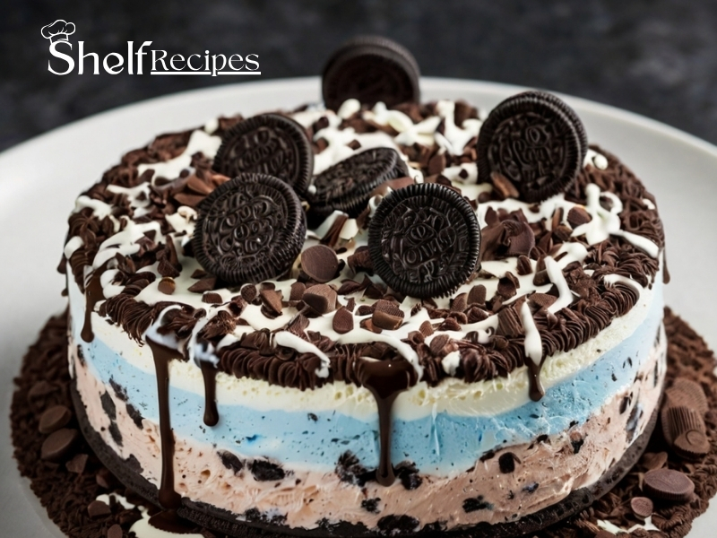 A layered Oreo ice cream cake with blue and pink ice cream layers, topped with chocolate drizzle, chocolate shavings, and whole Oreo cookies on a white plate. The cake is surrounded by a decorative ring of chocolate shavings.