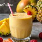 Glass of vibrant pineapple mango smoothie garnished with a pineapple slice and raspberry, surrounded by fresh pineapple, mango, strawberries, and raspberries on a grey surface.