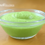 A clear glass bowl filled with a smooth, vibrant green wasabi sauce, placed on a wooden surface. The sauce has a slightly textured surface, indicating its fresh and creamy consistency.