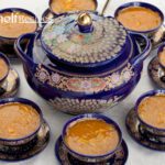 A beautifully arranged table setting featuring a large, ornate blue ceramic pot with intricate floral and peacock designs, filled with Moroccan Harira soup. Surrounding the pot are several matching blue bowls, each filled with the same rich, orange-colored soup, placed on decorative saucers. The setting is elegant and traditional, highlighting the cultural significance of the dish.