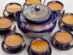 A beautifully arranged table setting featuring a large, ornate blue ceramic pot with intricate floral and peacock designs, filled with Moroccan Harira soup. Surrounding the pot are several matching blue bowls, each filled with the same rich, orange-colored soup, placed on decorative saucers. The setting is elegant and traditional, highlighting the cultural significance of the dish.