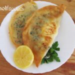 Two Tunisian brik pastries filled with egg and herbs on a white plate, garnished with a lemon wedge and parsley, on a wooden table.