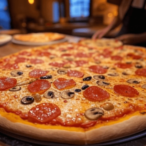 A close-up view of a freshly prepared pepperoni pizza, topped with sliced mushrooms and black olives. The pizza has a golden-brown crust and is placed on a metal tray. In the background, another pizza is visible, and a person appears to be working at a counter, possibly in a pizzeria or kitchen setting. The lighting is warm, suggesting a cozy, inviting atmosphere.