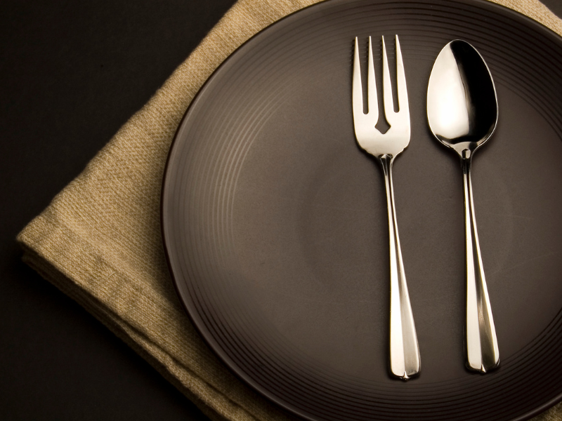 A neatly set empty plate with a fork and spoon on it, placed on a beige napkin against a dark background.