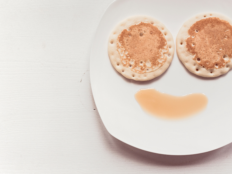 Two pancakes arranged on a white plate to resemble eyes, with syrup poured below them to form a smiling mouth.