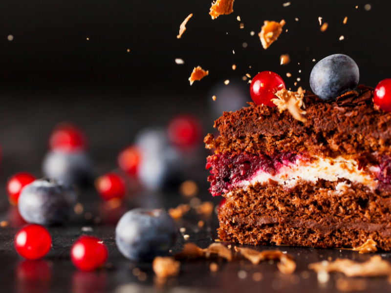 A close-up of a rich, layered chocolate dessert cake slice on a dark background, topped with blueberries and red currants, with crumbs flying around.