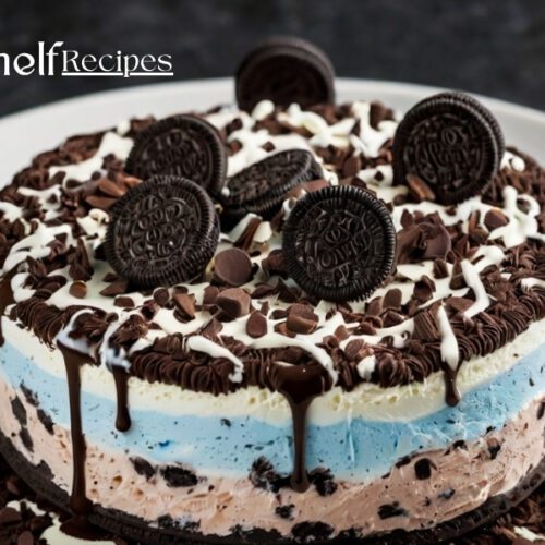 A layered Oreo ice cream cake with blue and pink ice cream layers, topped with chocolate drizzle, chocolate shavings, and whole Oreo cookies on a white plate. The cake is surrounded by a decorative ring of chocolate shavings.
